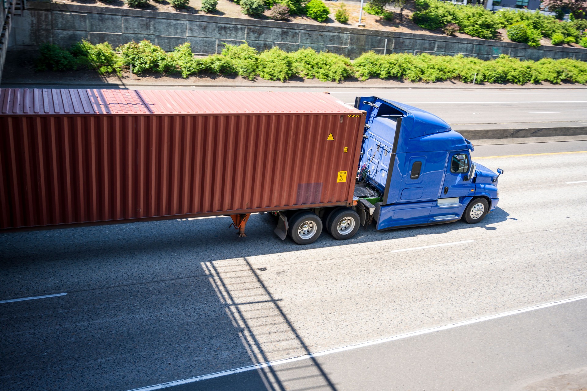 Blue comfort long haul big rig semi truck with long container driving on the divided highway road running under bridge